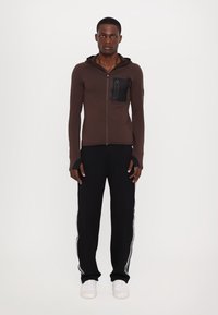 Ruda zip-up hoodie su tinkliniu kišenė, derinama su juodomis kelnėmis su baltomis šoninėmis juostelėmis ir baltomis sportinėmis bateliais. Bendras paviršius lygus.