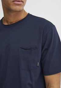 Tricou din bumbac de culoare bleumarin, cu guler rotund, mâneci scurte și un buzunar pe piept. Prezintă o etichetă mică pe cusătura de jos. Textură moale.