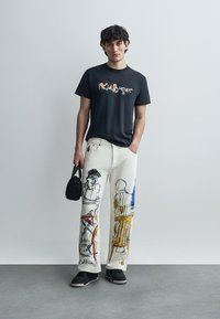 Μαύρο T-shirt με πολύχρωμο γραφικό σχέδιο, λευκό τζιν παντελόνι με καλλιτεχνικά σκίτσα, και μαύρα αθλητικά παπούτσια. Το μοντέλο κρατά μια μικρή μαύρη τσάντα.