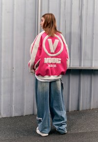 Chaqueta de satén rosa oversize con forro de felpa blanca, que presenta un logo circular y texto, combinada con pantalones de mezclilla azul holgados.