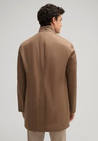 Lichte, bruin lange jas met een staande kraag. Heeft een rechte snit, geen zichtbare knopen en een rugventilatie. Zachte textuur en minimaal ontwerp.