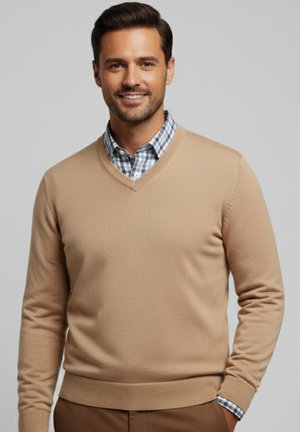 Hombre sonriente que lleva un suéter beige de cuello en V sobre una camisa a cuadros azul, de pie con una mano en el bolsillo frente a un fondo gris liso.