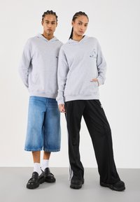 Šedé mikiny s předními kapsami, které nosí dva modelové. Jeden má na sobě denimové kalhoty typu culottes, druhý černé tepláky s bílými pruhy a robustními botami.
