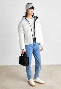 Giacca imbottita bianca con zip nera, fascia per capelli grigia in maglia, camicia bianca, jeans blu chiaro e sneakers bianche con accenti gialli. Borsa mini marrone.