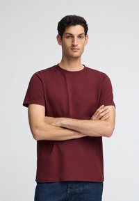 Bordeauxkleurig t-shirt met korte mouwen, gemaakt van zachte stof, met een ronde halslijn en een klein blauw logo-accent op de borst.