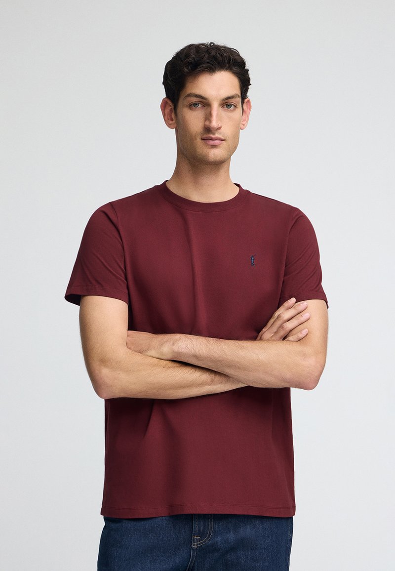 Bordeauxkleurig t-shirt met korte mouwen, gemaakt van zachte stof, met een ronde halslijn en een klein blauw logo-accent op de borst.