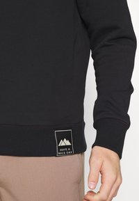 Sudadera negra con cuello y puños acanalados, que presenta una etiqueta rectangular con montañas y el texto "HAVE A NICE DAY" en blanco.