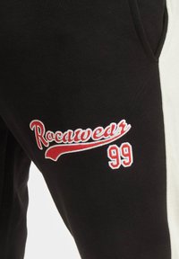 Juodos sportinės kelnės su baltomis šoninėmis juostelėmis, raudonai siuvinėtas tekstas "Rocawear" ir "99." Medžiaga atrodo minkšta su tekstūruotu paviršiumi.