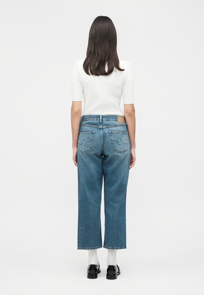 Γυναίκα που φαίνεται από πίσω, φοράει λευκό κοντομάνικο μπλουζάκι, μπλε ψηλόμεσο παντελόνι τζιν με φαρδιά μπατζάκια, λευκές κάλτσες και μαύρα παπούτσια σε απλό φόντο.