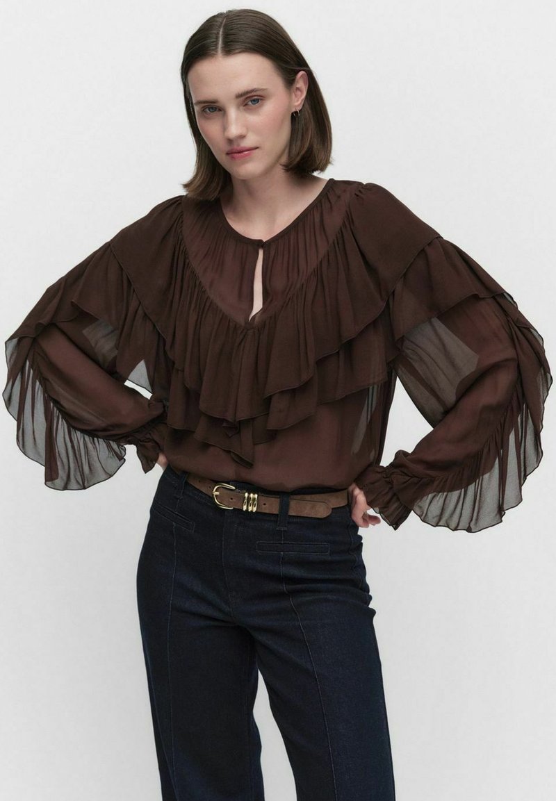 Blus chiffon cokelat dengan ruffles berlapis, detail leher kunci, dan lengan panjang mengalir. Dipadukan dengan celana jeans gelap dan sabuk.