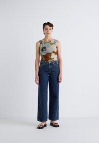 Top senza maniche, multicolore in tie-dye con pattern verdi e marroni, abbinato a jeans a gamba larga blu scuro a vita alta e sandali neri.