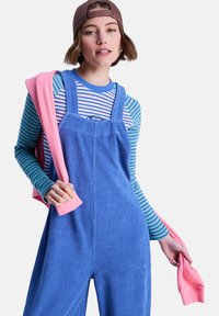 Μπλε βελούδινη σαλοπέτα με ρυθμιζόμενες τιράντες, φορεμένη πάνω από μια ριγέ μπλούζα μακρυμάνικη σε μπλε, πράσινο και λευκό, συμπληρωμένη με ροζ πουλόβερ.