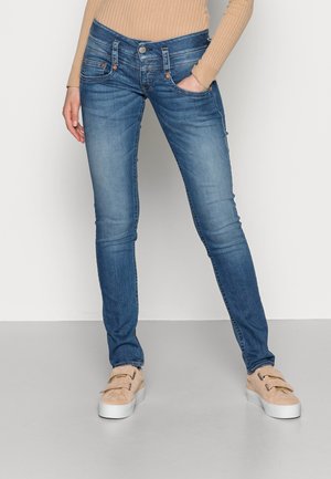 Frau trägt ausgewaschene blaue Skinny-Jeans und beige Slip-On-Sneaker, steht mit einer Hand in der Tasche vor einem einfarbigen Hintergrund.