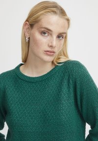 Maglione verde lavorato a maglia con un motivo testurizzato, scollatura a girocollo a coste e vestibilità rilassata. La modella ha lunghi capelli biondi e orecchini visibili.