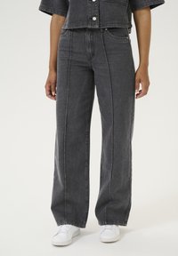 Wide-leg black denim jeans with contrasting stitching, high waist, and front pockets, paired with white sneakers. Smooth texture, classic design.