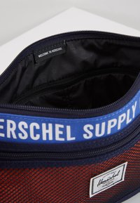 Příruční taška Herschel Supply s červenou síťovanou vnější částí, námořnickými detaily, modrou logovou páskou a černou vnitřní kapsou se štítkem "VÍTEJTE V HERSCHEL".