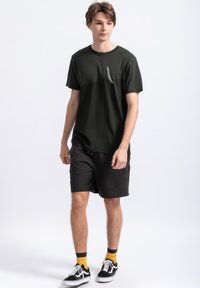 Donkergroene T-shirt met korte mouwen en een subtiele afbeelding op de borst, gecombineerd met zwarte shorts. Het model draagt zwarte sneakers met gele sokken.