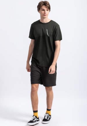 T-shirt verde scuro a maniche corte con una grafica sottile sul petto, abbinato a pantaloni corti neri. Il modello indossa scarpe da ginnastica nere con calzini gialli.