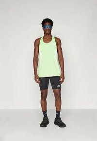 Grünes Sporttanktop mit weißen Akzenten, kombiniert mit schwarzen Shorts. Schwarze Turnschuhe und Sonnenbrille runden das Outfit ab.