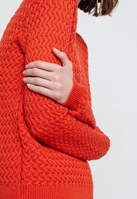Pull en tricot texturé de couleur orange vif. Caractérisé par des poignets à côtes épaisses et un motif ondulé sur l'ensemble du corps. Design ajusté.