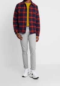 Camicia di flanella blu navy con una fantasia a quadri rossi e arancioni, abbinata a un pullover di maglia verde e pantaloni grigi chiari. Sneakers bianche con dettagli neri.