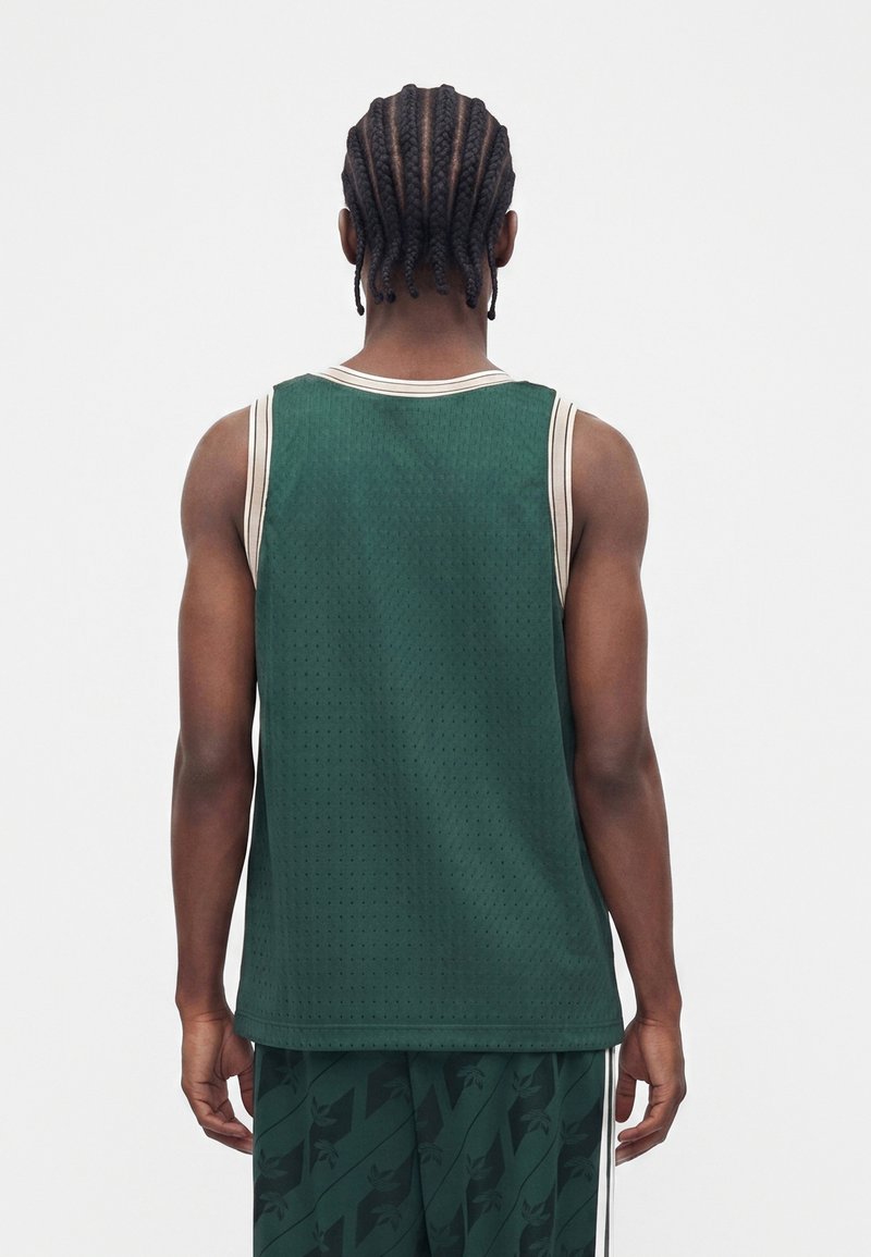 Mand med fletninger iført en mørkegrøn ærmeløs sportsvest og matchende mønstrede shorts, står med ryggen mod en hvid baggrund.