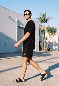 Mand iført sort t-shirt, shorts og sandaler går udendørs nær en hvid bygning med palmer i skarpt dagslys.