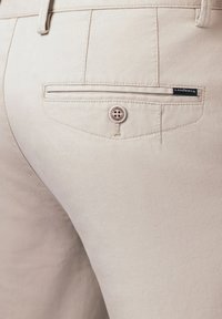 Zbliżenie tylnej kieszeni beżowych spodni z zapięciem na guzik oraz małą czarną metką Lancerto.