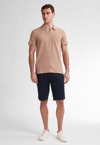 Beige lyhythihainen polo-paita taskulla, yhdistettynä tummansinisiin shortseihin. Yksivärisiä, sileällä pinnalla. Mallilla valkoiset tennarit.