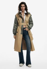Beige en olijfgroen dubbele trenchcoat met een tailleband, grote knopen, een kraag en zijzakken, gecombineerd met flared jeans.