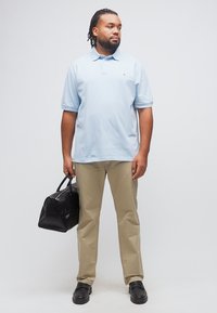 Άνδρας με πλεξούδες, φοράει ανοιχτό γαλάζιο πόλο και μπεζ παντελόνι, κρατώντας μια μαύρη τσάντα με υφή, κοιτάζοντας προς το πλάι.