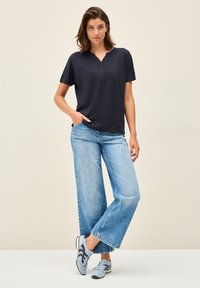Polo shirt bleu marine à manches courtes avec un col arrondi, associé à un jean en denim bleu clair à jambes larges et des baskets bleues, se tenant devant un fond neutre.