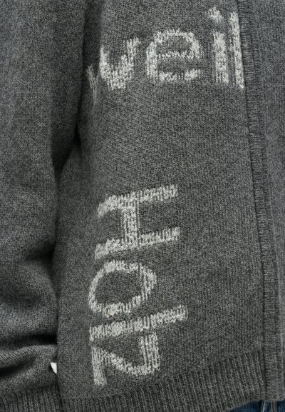 Pilkas megztinis su kontrastine šviesiai pilka užrašu. Minkšta tekstūra su matomais siūlais ir šoninės siūlės detale.