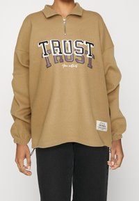 Person iført en beige halvzip-pullover med teksten "TRUST" og "Your instincts" samt sorte bukser, stående med hænderne afslappet ned langs siden.