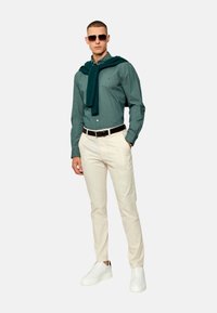 Camisa verde con estampado, pantalones blancos, zapatillas blancas y un suéter verde azulado caído sobre los hombros. Las gafas de sol completan el look.