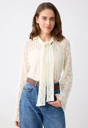 Femme portant un chemisier en dentelle crème transparente avec un nœud noué au cou et un jean bleu taille haute ceinturé de marron.