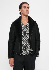 Joven vestido con una chaqueta de borreguito negra sobre una camisa de botones con estampado en blanco y negro y pantalones negros, mirando ligeramente hacia arriba.