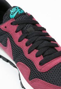 Svart och mörkröd Nike Air sneakers med teal logotyp, svarta snörningar, nät- och mocka-texturerade paneler, visad från en vinklad närbild.