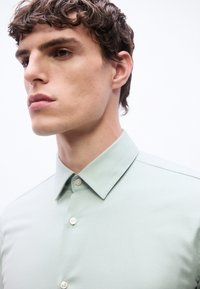 Νεαρός με κοντά σγουρά μαλλιά που φοράει ανοιχτοπράσινο πουκάμισο με γιακά, στραμμένος προς τα αριστερά σε λευκό φόντο.