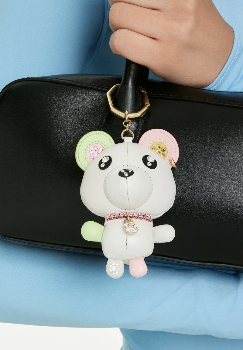 Hand håller en svart läderhandväska med en vit björnnyckelring prydd med pastellfärgade öron, rosa halsband och glittrande ädelstensdekorationer.