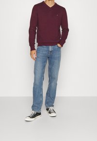Mężczyzna ubrany w burgundowy sweter w serek, jasnoniebieskie dżinsy i czarno-białe sneakersy, stojący z jedną ręką w kieszeni.