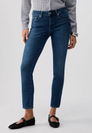 Donna che indossa jeans skinny blu scuro a vita media, maglione grigio a trecce, ballerine nere Mary Jane e braccialetti d'argento al polso destro.