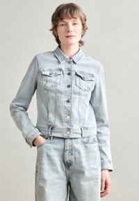Svijetloplavi traper jakna s ovratnikom, prednjim džepovima s preklopima, dva džepa na prsima i kopčanjem na gumbe. Ima usku struk i duge rukave.