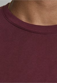 Gros plan sur un cou portant une chaîne en or et un t-shirt à col rond bordeaux, montrant la texture du tissu et les détails de la peau.