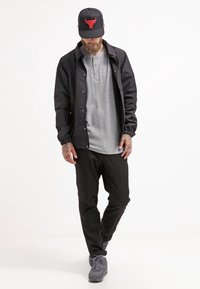 Man i svart jacka, grå henley-tröja, svarta byxor, grå sneakers och svart keps med Red Bull-logga, stående med huvudet nedåt.