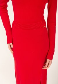 Rotes Strickkleid mit langen Ärmeln und einem gerippten Band an der Taille. Verfügt über einen seitlichen Schlitz und ein kleines goldenes Akzentdetail. Glatte Textur.