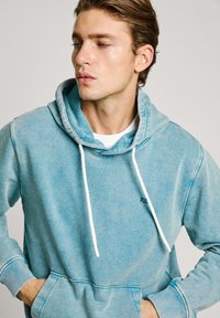 Lichtblauwe hoodie van een zachte stof met een kangoeroezak, witte trekkoorden en ribbelmanchetten; heeft een subtiele logo op de borst.