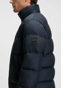 Marinblått pufferjacka med hög krage, quiltad textur och en liten svart logotyp på ärmen. Mjukt, lätt material.