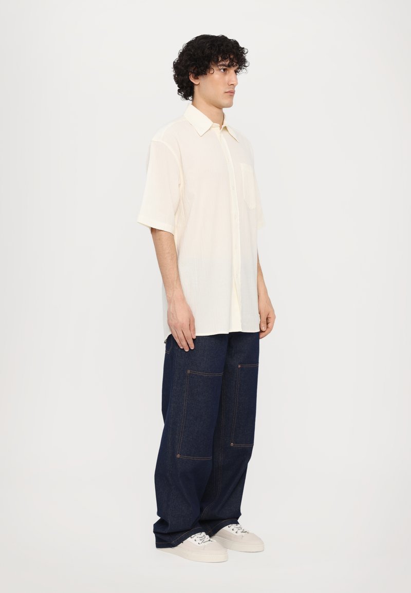 Camisa de manga corta color crema con cuello tipo botón, combinada con pantalones de mezclilla azules de corte holgado que tienen grandes bolsillos y dobladillos en los extremos.