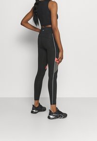 Černé legíny s texturovaným vzorem, vysokým pasem, stříbrným bočním zipem a růžovými kapsami. V páru s černými sportovními botami.